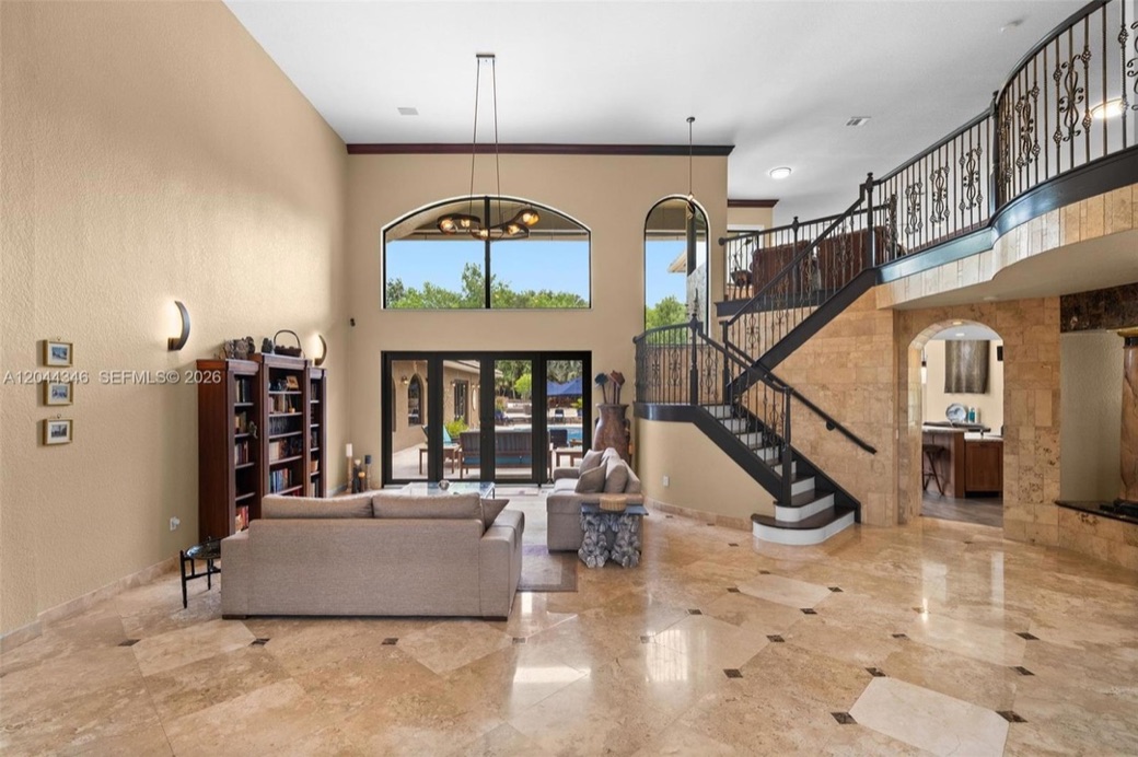 Grand two-story living room with a curved wrought-iron staircase, marble floors and pool views through floor-to-ceiling glass